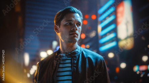 Portrait of Handsome Serious Man Standing, Looking Around Night City with Bokeh Neon Street Lights in Background. Focused Confident Young Man Thinking. Portrait Shot.