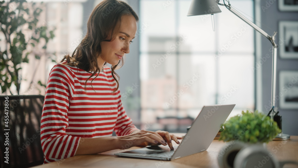 Young Woman Using Laptop at Home for Remote Work. Beautiful Young Brunette Girl Sitting at Her Desk Uses Internet, Doing Productive Distant Work. Side View Shot Young Woman Using Laptop at Home for Remote Work. Beautiful Young Brunette Girl Sitting at Her Desk Uses Internet, Doing Productive Distant Work. Side View Shot