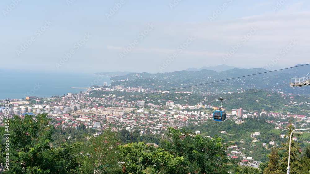 Obraz premium Cable car cabin over green trees and city houses with mountain and sea views in the background on a summer sunny day
