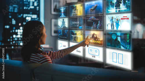 Bild auf Leinwand Beautiful Young Woman is Sitting on a Couch at Night at Home and Choosing a Movie to Watch on a Futuristic Augmented Reality Hologram Screen