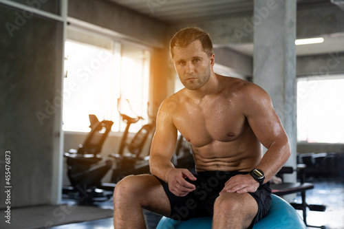 Muscular man sitting on ball and take a break after his workout in the fitness gym