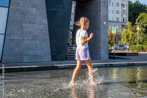 фотографія A little girl of 8 years old runs in the city fountain and splashes against the