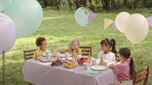 High-angle of four diverse gradeschoolers sitting at festive table in sunny park, having birthday party, talking photo