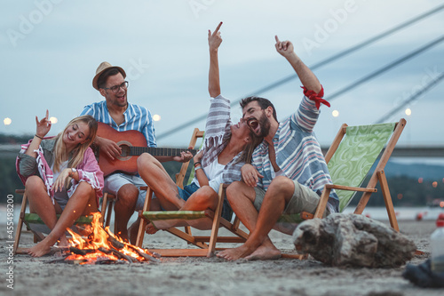 On a tranquil autumn evening, a group of friends gathers by the riverside sandy beach. They sit around a small campfire, under the starlit sky, enjoying their drinks, singing, and playing guitar. © BalanceFormCreative