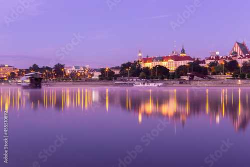 Illuminated Warsaw Old Town landscape with the Royal Castle, Cathedral and medieval buildings, during purple sunrise with reflections in calm Vistula river, Poland.