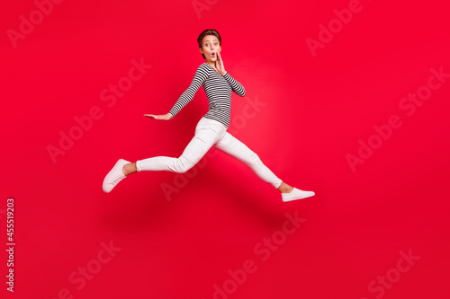 Photo of shiny impressed young woman dressed striped outfit arm cheek jumping hign isolated red color background