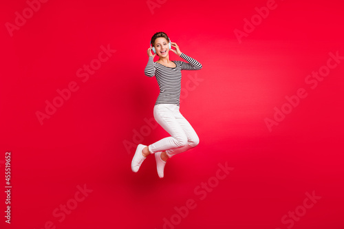 Photo of shiny funny young lady wear striped clothes headphones smiling jumping high isolated red color background
