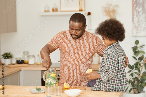 African man mixing lemonade in jug while his little son with fresh lemon half in hand looking at him