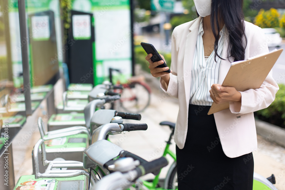 Asian business woman wearing protective face mask prevent covid-19 virus using smartphone for rent public bicycle outdoors. Bicycles for rent in city environment. Asian business woman wearing protective face mask prevent covid-19 virus using smartphone for rent public bicycle outdoors. Bicycles for rent in city environment.
