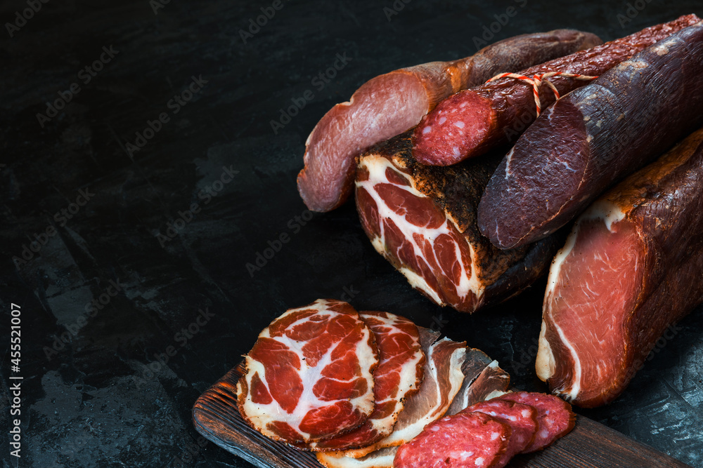 Fototapeta premium Whole and sliced meat of ham, sausage, prosciutto, jamon, bacon. Meat antipasto, banner, menu, black background. Copy space.