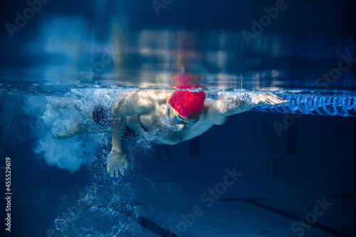 Underwater view of professional male swimmer in red cap and goggles in motion and action during training at pool, indoors. Healthy lifestyle, power, energy, sports movement concept