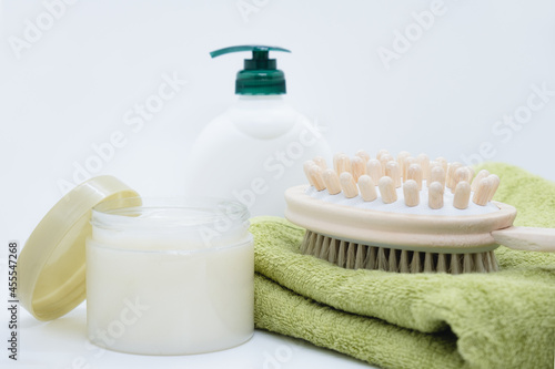 Double-sided massage brush for body brushing lies on green bamboo towel on white background. Dispenser cream and open jar of anti-cellulite body scrub. Materials for spa treatments