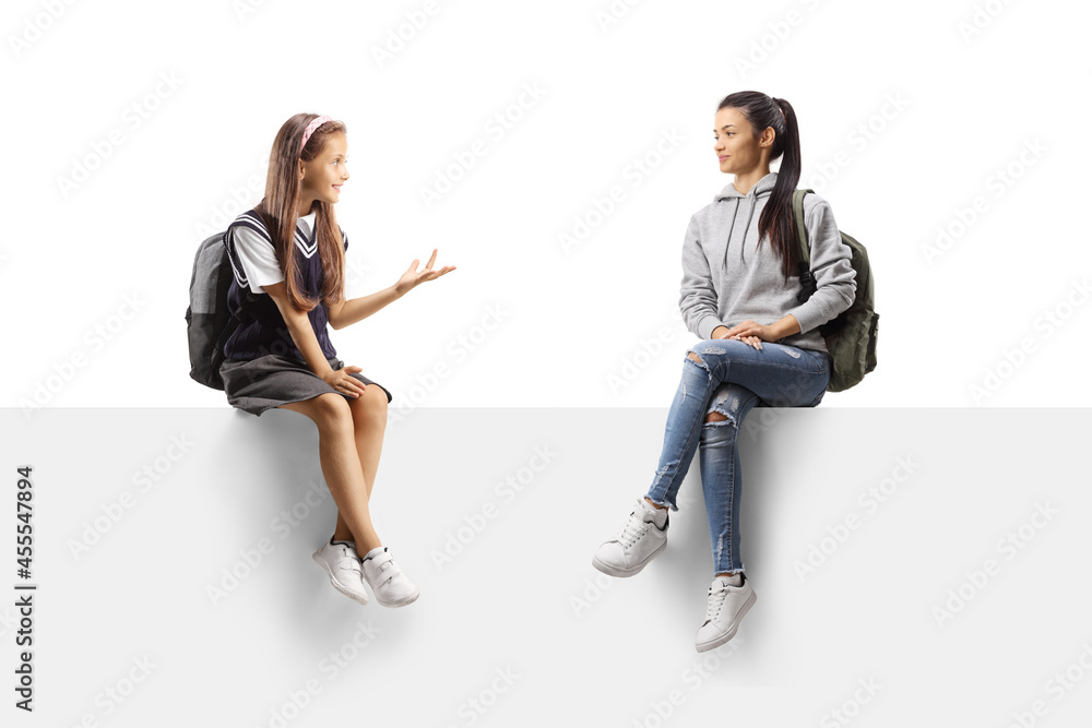Little schoolgirl talking to a young female student and sitting on a blank panel