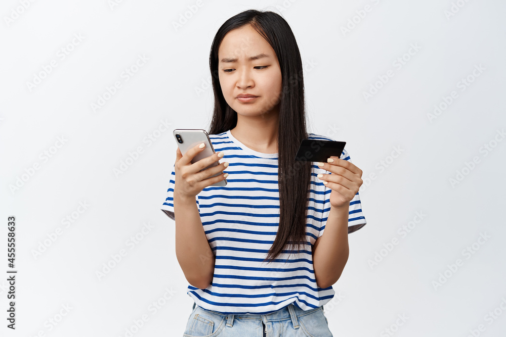Asian girl has difficulties with online shopping app, problem during payment, frowning at mobile phone screen, holding credit card, standing over white background Asian girl has difficulties with online shopping app, problem during payment, frowning at mobile phone screen, holding credit card, standing over white background