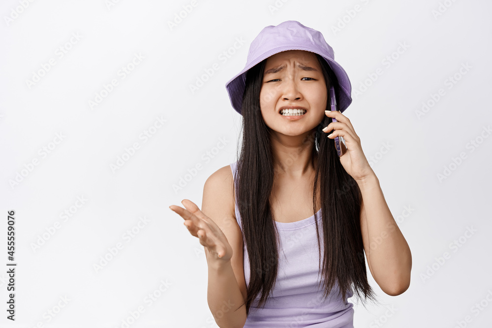 Upset asian girl looking troubled while having a phone call, frowning and staring distressed, having upsetting conversation, white background Upset asian girl looking troubled while having a phone call, frowning and staring distressed, having upsetting conversation, white background