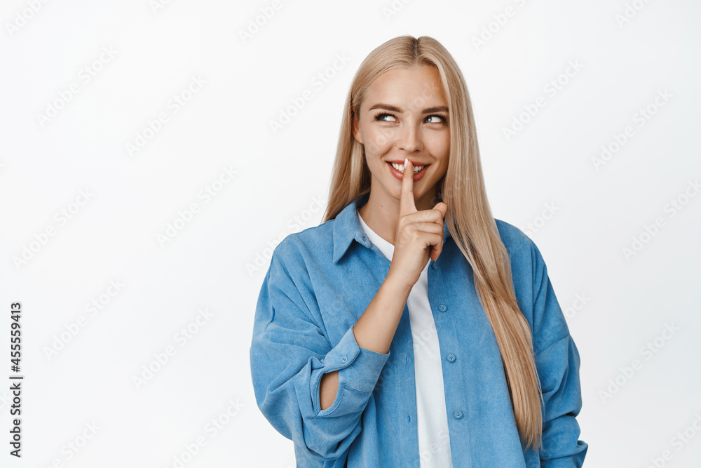 Mysterious blond girl hushing and smiling, looking at upper left corner product and shushing, making shh taboo gesture, standing over white background Mysterious blond girl hushing and smiling, looking at upper left corner product and shushing, making shh taboo gesture, standing over white background