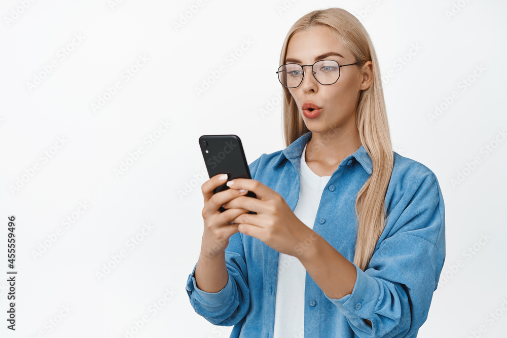 Portrait of young female student in glasses looking at her mobile phone with curious face expression, reading message on smartphone, white background Portrait of young female student in glasses looking at her mobile phone with curious face expression, reading message on smartphone, white background