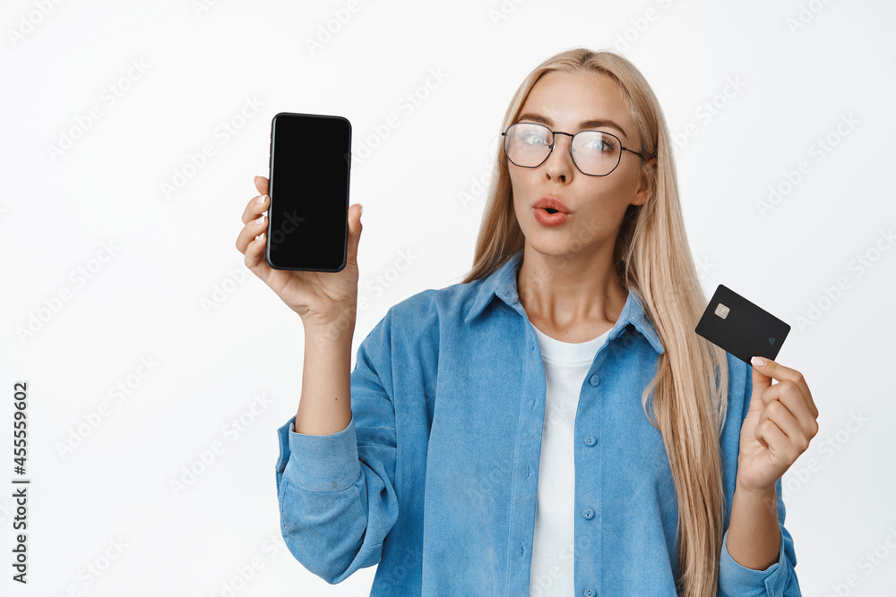 Portrait of woman in glasses looking curious, showing interesting app on mobile phone and credit card, concept of online shopping and contactless payment, white background Portrait of woman in glasses looking curious, showing interesting app on mobile phone and credit card, concept of online shopping and contactless payment, white background