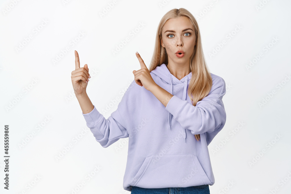 Curious blond girl pointing fingers at upper left corner, looking intrigued, asking question of advertisement, standing over white background Curious blond girl pointing fingers at upper left corner, looking intrigued, asking question of advertisement, standing over white background