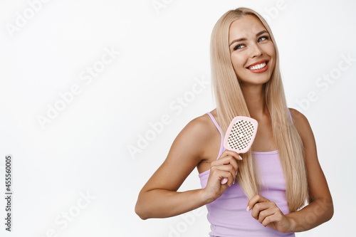 Good-looking young blond woman combing hair, holding comb brush and smiling, looking aside, standing in tank top over white background