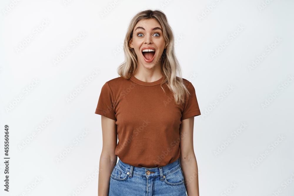 Fototapeta premium Image of blond girl looking surprised and excited, open mouth and widen eyes impressed, standing in t-shirt and jeans over white background
