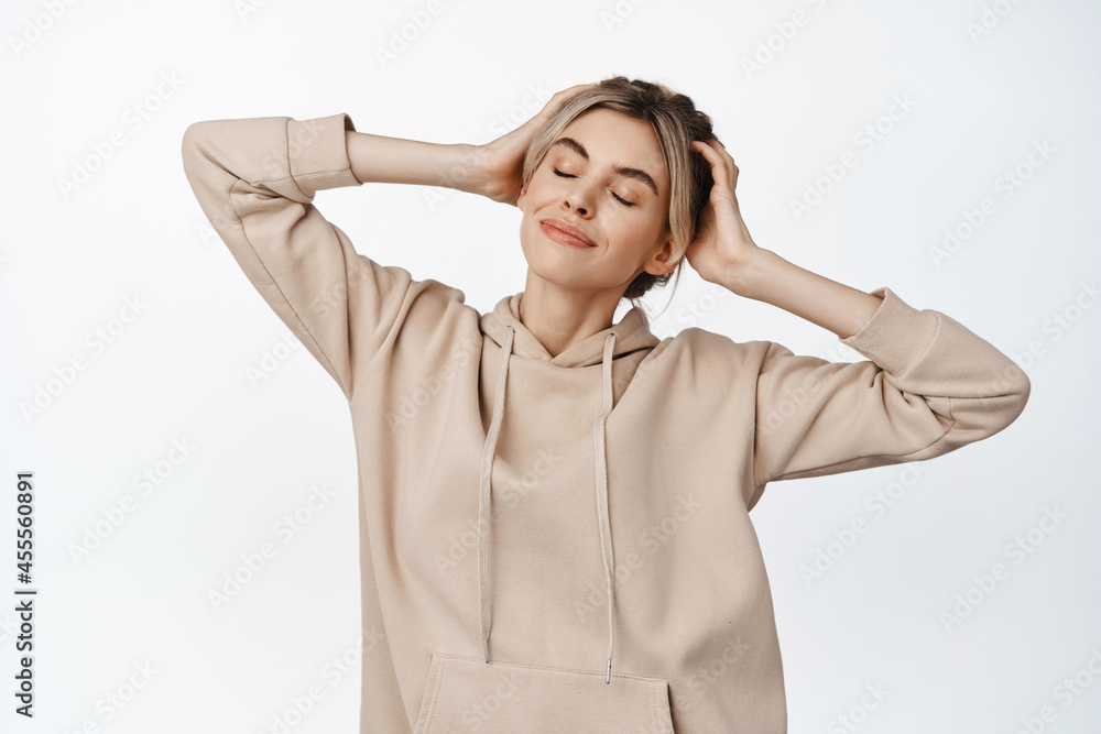 Tender and beautiful blond girl close eyes, smiling and touching her hair with satisfaction, standing in beige hoodie over white background Tender and beautiful blond girl close eyes, smiling and touching her hair with satisfaction, standing in beige hoodie over white background