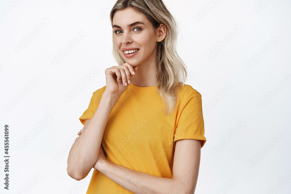 Portrait of natural young woman with blond hair, smiling and looking thoughtful at camera, making decision, standing in yellow t-shirt against white background Portrait of natural young woman with blond hair, smiling and looking thoughtful at camera, making decision, standing in yellow t-shirt against white background