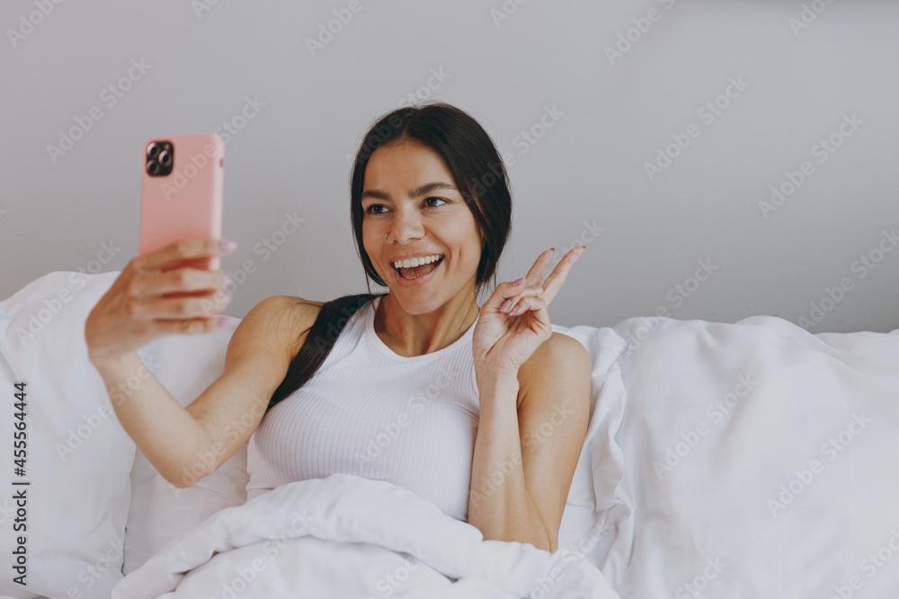Excited young woman in white tank top sit in bed doing selfie shot on mobile cell phone post photo on social network in bedroom lounge home in own room house wake up dream be lost in reverie good day.
