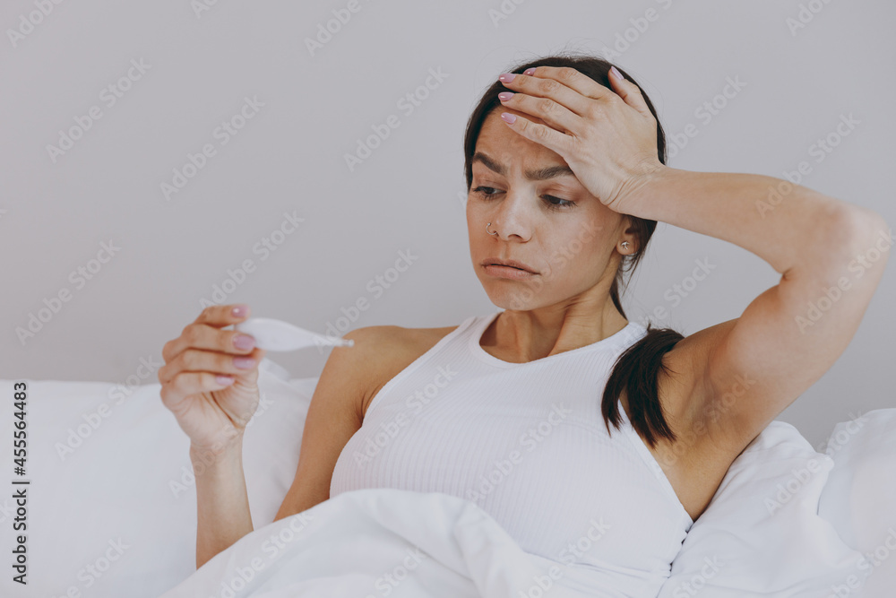 Sick ill tired sad young woman in white tank top sit in bed hold hand on forehead thermometer in bedroom lounge home in own room house wake up. Healthy lifestyle disease treatment cold season concept. Sick ill tired sad young woman in white tank top sit in bed hold hand on forehead thermometer in bedroom lounge home in own room house wake up. Healthy lifestyle disease treatment cold season concept.