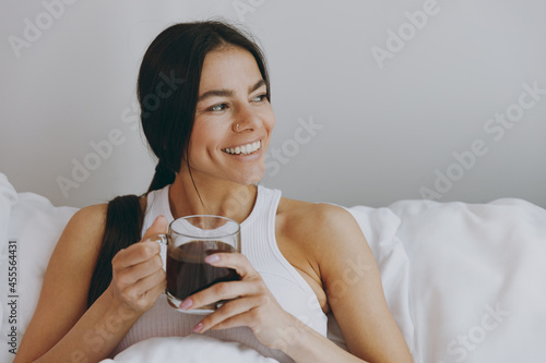 Calm happy young woman in white tank top sit in bed hold drink coffee from transparent glass rest relax spend time in bedroom lounge home in own room house wake up dream be lost in reverie good day. photo