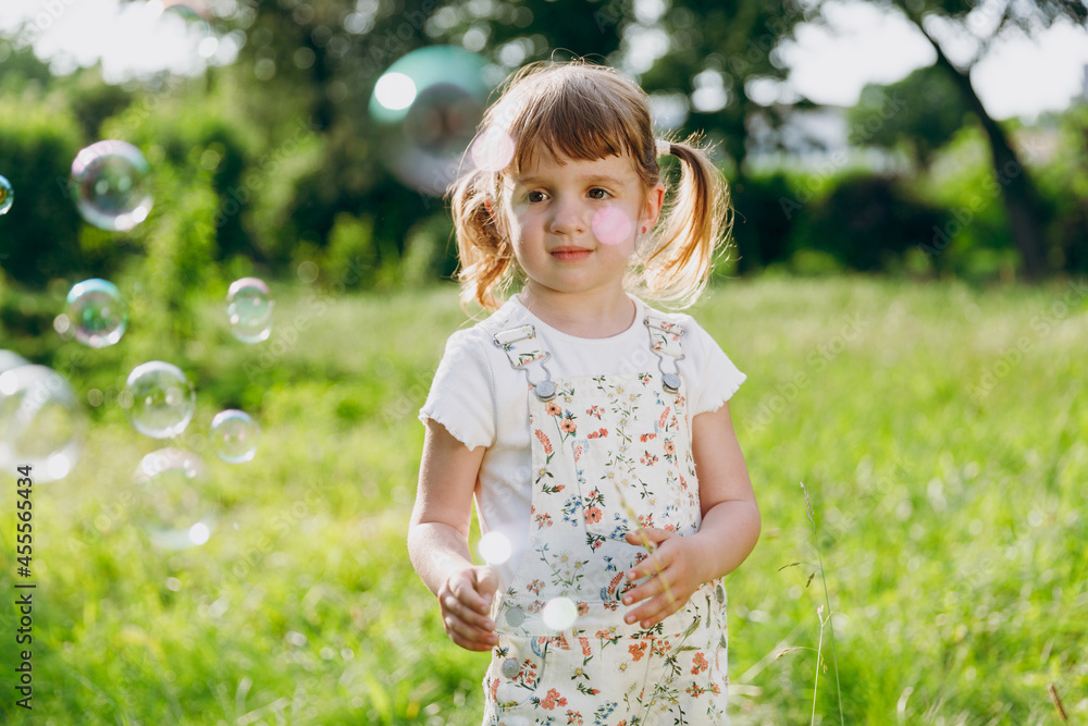 Little smiling satisfied happy kid girl 5-6 years old in white casual clothes blowing bubbles play on park green sunshine lawn, spending time outdoor in village countyside during summer time vacation Little smiling satisfied happy kid girl 5-6 years old in white casual clothes blowing bubbles play on park green sunshine lawn, spending time outdoor in village countyside during summer time vacation