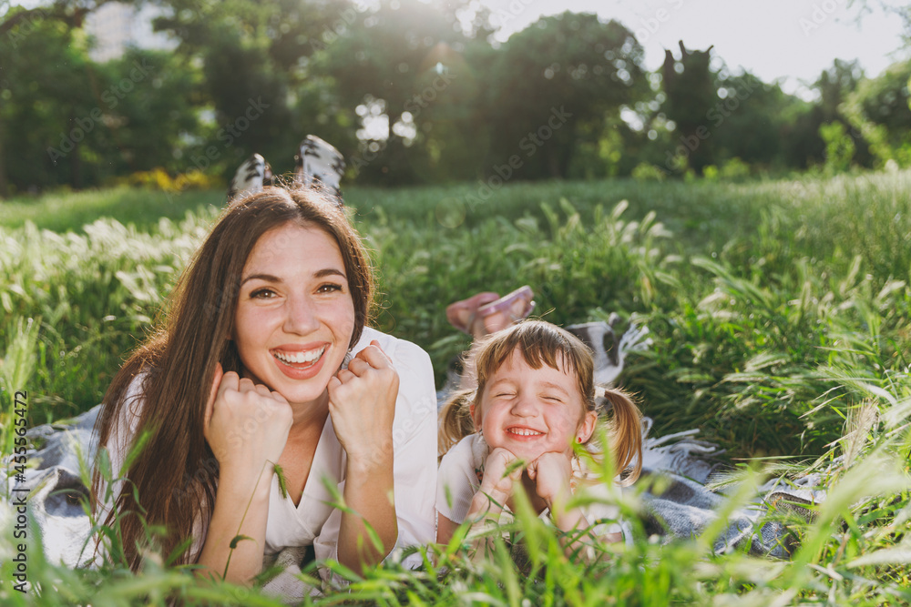 Fototapeta premium Happy satisfied young woman in white clothes have fun picnic lying on plaid grass with child baby girl 5-6 years old. Mommy play rest with little kid daughter outdoor together. Love family concept