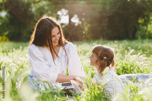 Happy smiling sunlit young woman in white clothes have fun picnic sit on plaid grass with cute child baby girl 5-6 years old. Mommy rest with little kid daughter outdoor together. Love family concept