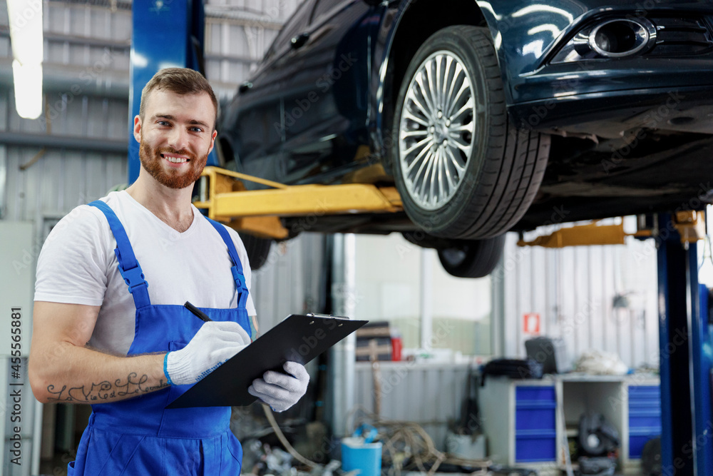 Young happy technician mechanic man wears denim overalls use hold clipboard papers document writing estimate stand near car lift check technical condition work in vehicle repair shop workshop indoors.