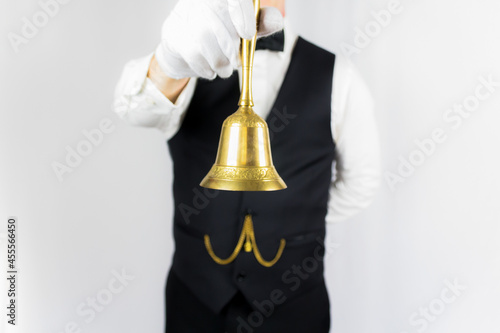 Portrait of Butler or Waiter in Black Vest and Bow Tie Holding Gold Bell on White Background. Concept of Service Industry and Professional Hospitality. 