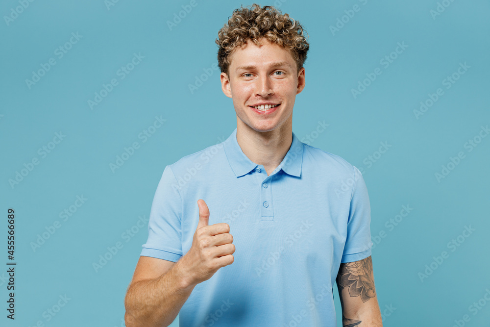 Smling young curly man 20s years old wear azure t-shirt looking camera showing thumbs up like gesture isolated on plain pastel light blue background studio portrait. People emotions lifestyle concept Smling young curly man 20s years old wear azure t-shirt looking camera showing thumbs up like gesture isolated on plain pastel light blue background studio portrait. People emotions lifestyle concept