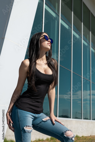 young woman with long black hair  slim posing standing by a building  wearing casual blouse  fashionable sunglasses and jeans  beauty and modern lifestyle