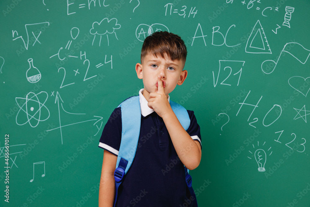 Young male kid school boy 5-6 year old in t-shirt backpack say hush be quiet with finger on lips isolated on green wall chalk blackboard background Childhood children kids education lifestyle concept.