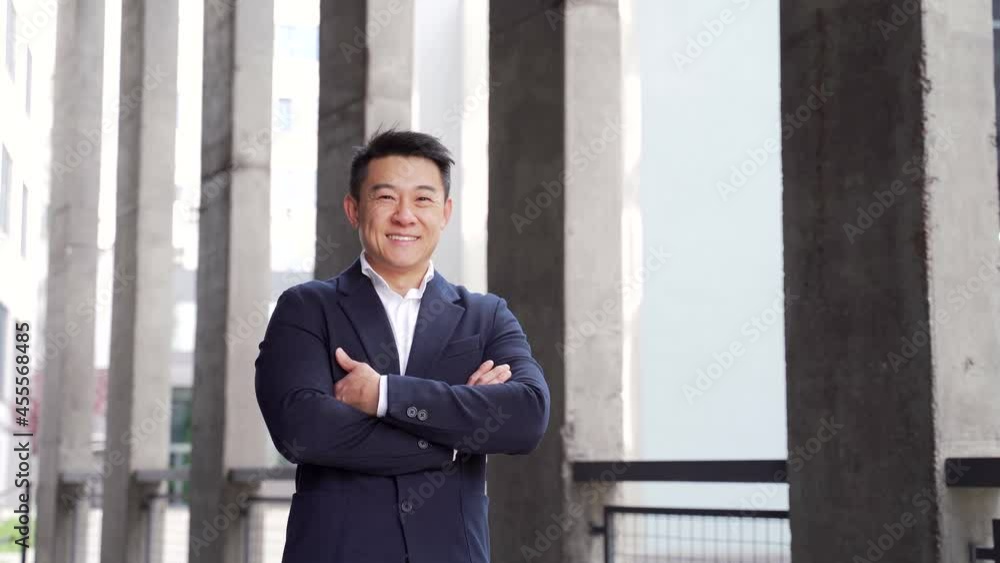 Portrait happy friendly asian businessman standing on urban city street background outside. Business man office worker arms crossed in formal suit near modern office building looking at camera smiling Portrait happy friendly asian businessman standing on urban city street background outside. Business man office worker arms crossed in formal suit near modern office building looking at camera smiling