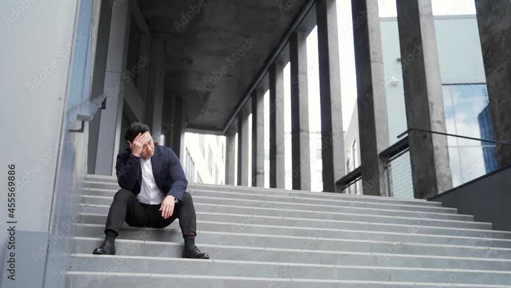 young asian depressed man sitting on the stairs outdoors street near office business center building. Upset male businessman lost his job due financial crisis and quarantine. Businessman has problems young asian depressed man sitting on the stairs outdoors street near office business center building. Upset male businessman lost his job due financial crisis and quarantine. Businessman has problems