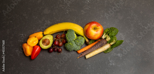 Fruit and vegetable arrangement on gray granite surface including bananna, apple, grapes, broccoli, avacado, carrots, spinich and bell peppers