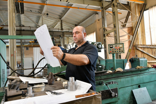 A worker reads a technical drawing near a metalworking machine before starting work.