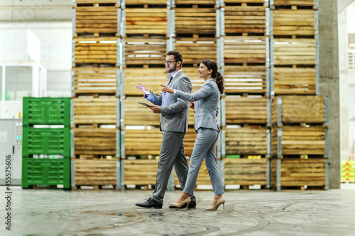Warehouse Management. A man and a woman in a business suit enter the warehouse and advance the business. The man holds the tablet in his hand and points his finger while the woman imitate