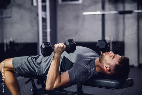 An attractive and muscular man prepares to lift weights. A man in sportswear pumps his arm muscles and lifts dumbbells in a modern gym. A close-up shot of a male person lying on a sports bench. Sport © dusanpetkovic1