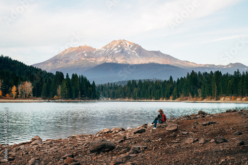 Person with fedora hat and back bag sitting on river shore in woods near near mountain photo