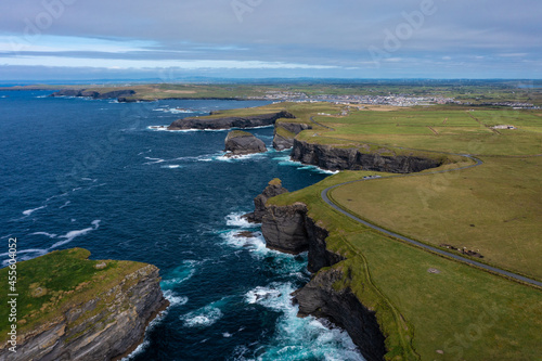 Aerial view of beautiful and green mountain of the famous tourist landmark Moher Cliff in Ireland with sea waves hitting rocks during day under cloudy sky