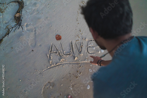 Man writes with his finger the word alive on the sandy seashore with water washing over the letters, top view