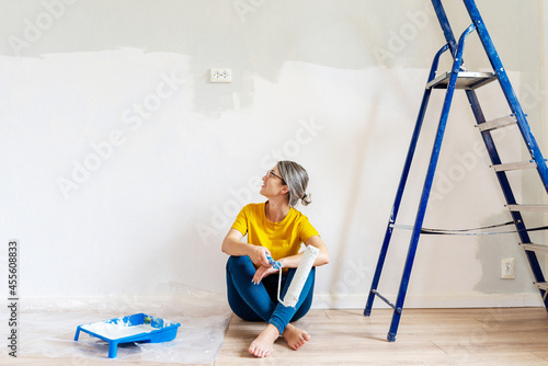 A woman in a yellow T-shirt is painting a wall in her house. A girl sits next to a painted wall, next to a stepladder