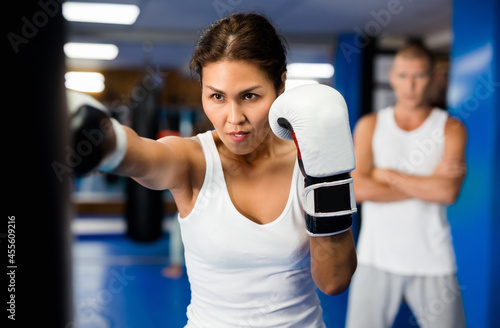 Asian woman in boxing gloves exercising jabs with punchbag during her boxing training. Her trainer standing behind and observing.