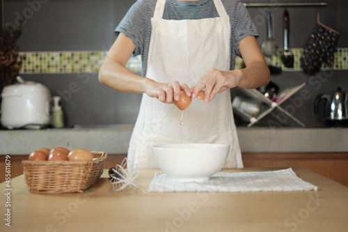 Close-up front view footage, a female cook in a white apron is cracking an egg into a cup to prepare a meal on a wooden table in the home's kitchen. Eating egg yolks is a healthy breakfast. photo